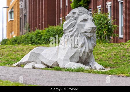 Modern geometric lion sculpture made of concrete, situated on a grassy area near a sidewalk in an urban neighborhood with brick and wooden buildings. Stock Photo