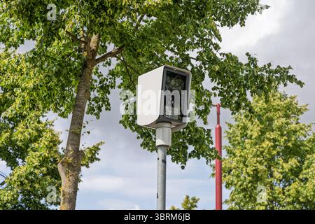 A modern white surveillance speed camera (flitspaal) mounted on a pole amidst green trees and a bright sky, ensuring safety and monitoring. Stock Photo