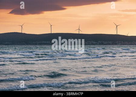 Wind farm generating renewable energy at sunset, casting a warm glow over the wavy sea Stock Photo