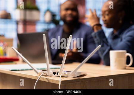 Frustrated African American woman tries to reset the Wi Fi modem while her partner checks laptop settings, troubleshooting network errors affecting their online work and browsing experience. Stock Photo