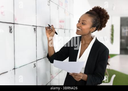 Smiling African businesswoman holding papers, writing on whiteboard Stock Photo