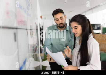 Teammates collaborating on paperwork standing next to large white board Stock Photo
