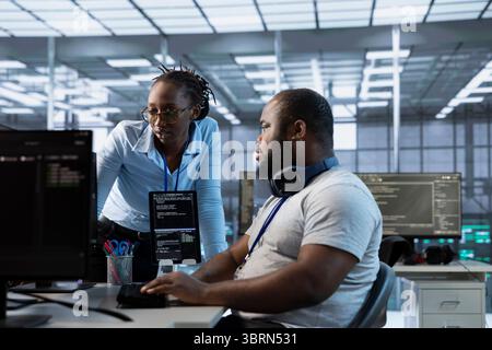 Technician requesting help from manager while working on computer in data center, monitoring hardware energy consumption. Team leader overseeing IT specialist checking for system failures Stock Photo