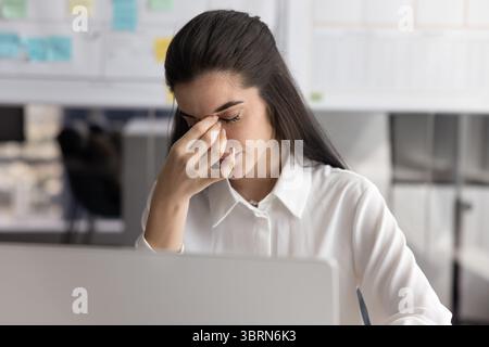 Woman sits at desk with laptop, closing eyes, feels tired Stock Photo