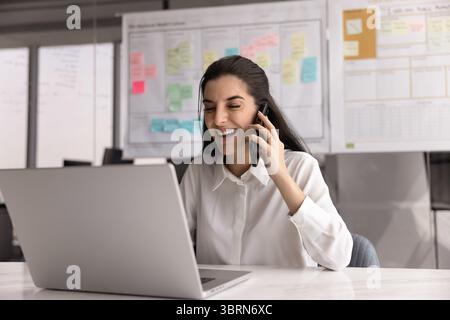 Laughing woman talking on smartphone using laptop working in workplace Stock Photo