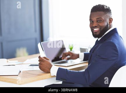 Cheerful black general manager checking reports, sitting in office Stock Photo