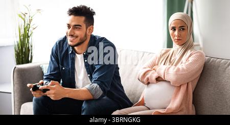 Upset Expectant Muslim Woman Sitting Offended To Husband That Playing Video Games Stock Photo