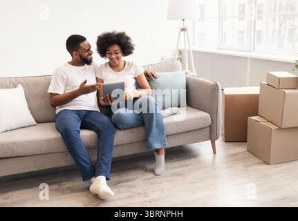 Happy Black Spouses Using Tablet Sitting Among Moving Boxes Indoor Stock Photo