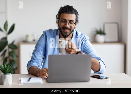 Young Smiling Arab Freelancer Guy Using Laptop And Drinking Coffee At Home Stock Photo