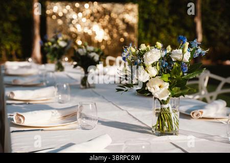 Outdoor dining table elegantly set with white linens and glassware, adorned with fresh floral centerpieces. Blurred golden lights in the background cr Stock Photo