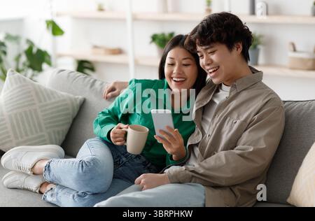 Positive young Asian couple using smartphone and drinking coffee while resting on couch at home, free space Stock Photo