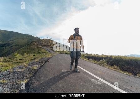 Hiker in yellow jacket holding a phone while walking alone on a hilly road. Stock Photo