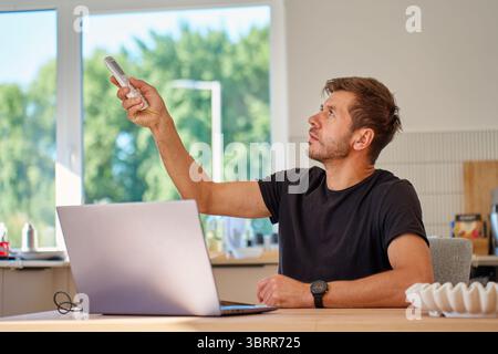Man sitting at desk with laptop and using remote control to adjust temperature for cooling at hot weather Stock Photo