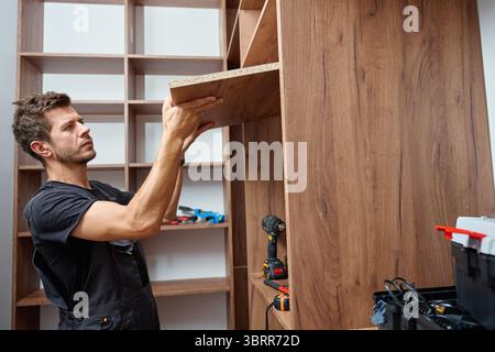 Man assembling wooden wardrobe at home using tools and holding large wooden panel. Concept of DIY furniture assembly, home improvement and renovation Stock Photo