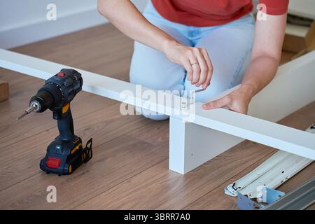 Woman assembling bed frame on wooden floor, using wrench and drill for installation. DIY furniture assembly and home improvement Stock Photo
