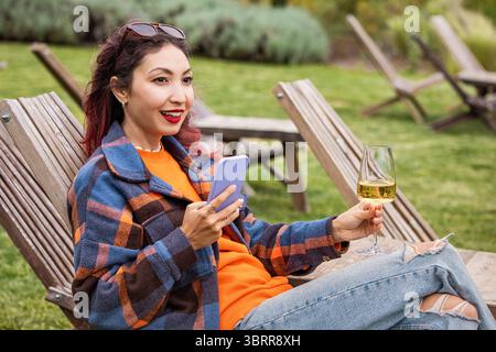 Tourist enjoying a glass of wine while using her smartphone, sitting on a wooden deck chair in a winery garden Stock Photo