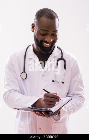 Portrait of cheerful black male paramedic in medical uniform who is writing notes in personal organizer. Studio shot on gray background. Stock Photo