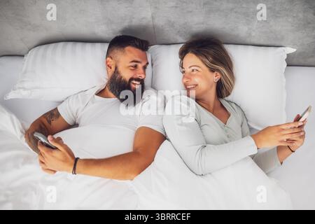 Young happy couple is lying in bed in the morning. They are relaxing and using smartphones. Stock Photo