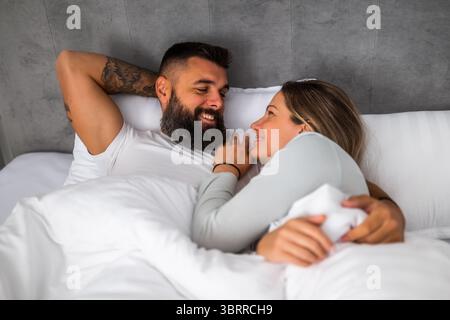 Young happy loving couple is lying in bed in the morning. They are relaxing and enjoying. Stock Photo