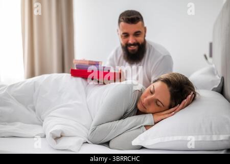 Young couple is lying in bed in the morning. Man is giving presents to his wife for her birthday. Stock Photo