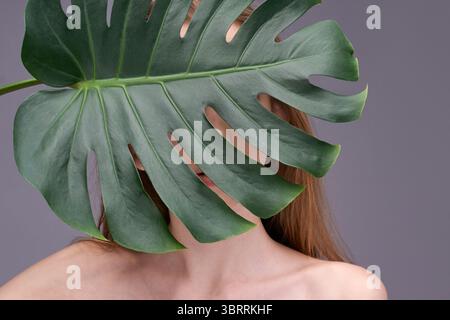 Mysterious young caucasian woman partially hidden behind large monstera leaf against gray background. Stock Photo