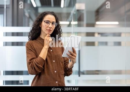 A thoughtful woman in glasses, holding a tablet, considers something indoors with glass walls. Stock Photo