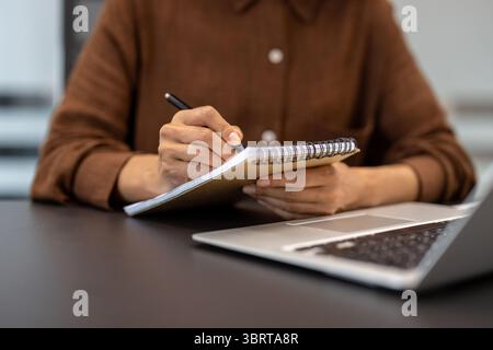 A person writes in a spiral notebook with a pen, with a laptop visible in the background, capturing productivity and note-taking. Stock Photo