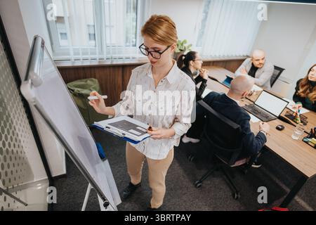 Team of colleagues collaborating during a business meeting in a modern office environment with focus on a woman presenting at a whiteboard Stock Photo