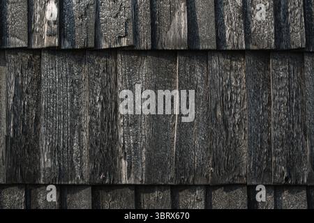 A detailed close-up of a rustic, dark, weathered, and charred black wooden shingle wall, showcasing a rough textured surface background. Stock Photo