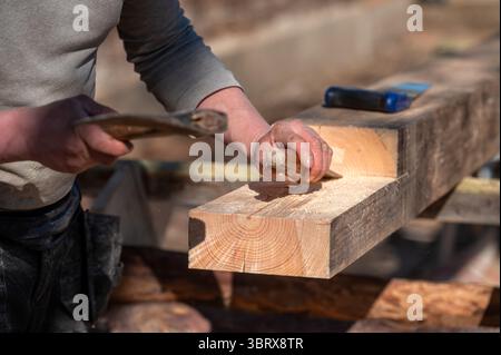 A carpenter skillfully shapes a wooden beam using a chisel and hammer in sunlight. Stock Photo