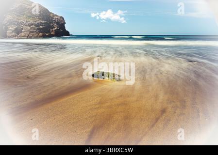 A long exposure photograph captures the ethereal flow of waves washing over a sandy beach, blurring the water in Portreath, Cornwall, UK Stock Photo