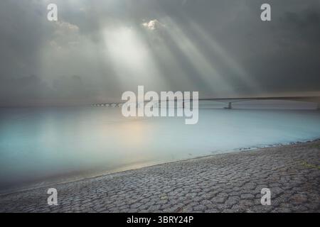 Long exposure image captures the Zeeland Bridge extending over calm waters, beautifully illuminated by dramatic sunrays breaking through a cloudy sky. Stock Photo