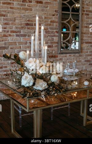 A sophisticated candlelit floral centerpiece creates an inviting ambiance on a glass table in a rustic setting, featuring white roses and gold leaves Stock Photo