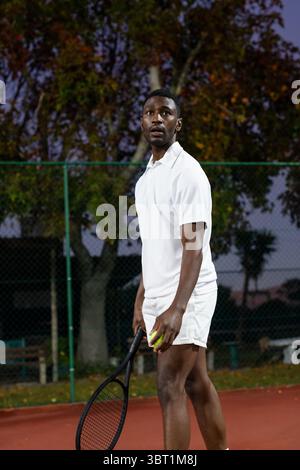 African American man wearing white tennis attire holding racket and ball on red court at dusk Stock Photo