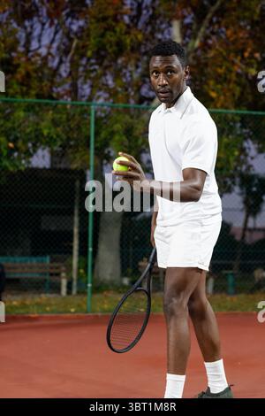 African American man standing on red park court holding racket and ball near green chain-link fence Stock Photo