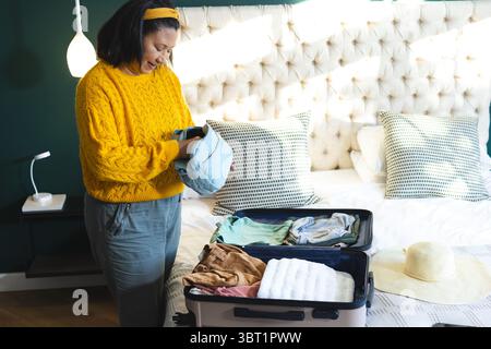 Middle-aged woman holding folded clothing and packing suitcase on bed in room with tufted headboard Stock Photo