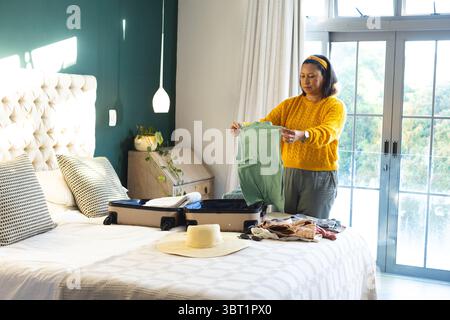 Woman packing clothes into suitcase on bed in bedroom under pendant lamp by window, copy space Stock Photo