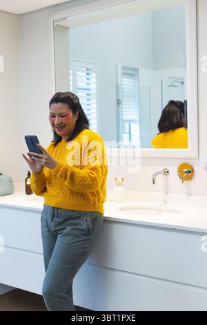 Woman in her 40s standing at sink in bathroom using smartphone with eye patches, copy space Stock Photo