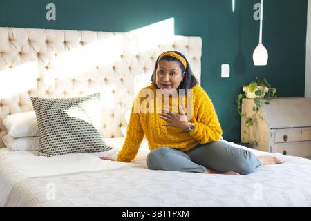 Woman speaking while sitting cross-legged on bed in bedroom with tufted headboard and smartwatch Stock Photo