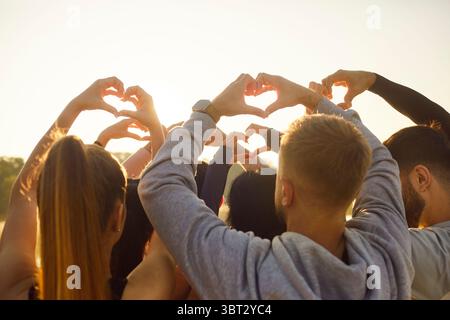 Friends group making heart shape gesture with fingers in sunlight, people raise hands to sun Stock Photo
