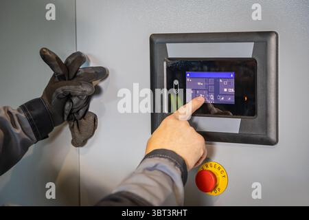 Electrician interacting with touchscreen of modern control panel, managing industrial processes and ensuring safety with emergency stop button Stock Photo