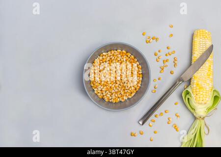 Corn grains in gray bowl. Knife lies on cob of corn. Gray background. Flat lay Stock Photo