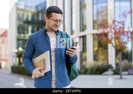 A young man in glasses is using his phone outside a modern building, carrying a laptop and notebook. Stock Photo