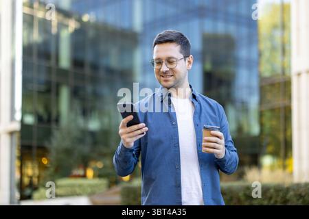 A man in glasses smiles while looking at his phone, holding a coffee cup in front of a modern building. Stock Photo