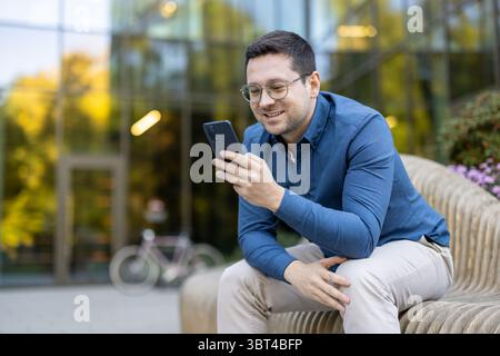A smiling man in glasses checks his phone while sitting on a bench outdoors near an office building. Stock Photo