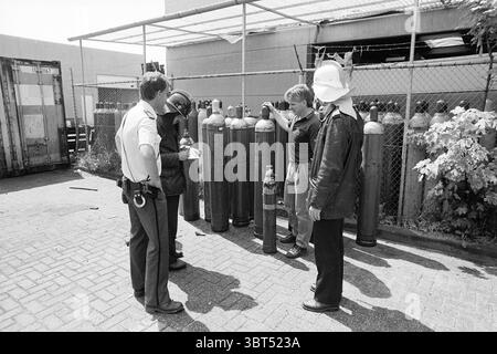 Explosion of carbon dioxide bottle company R. Peereboomweg Explosions Haarlem Robert Peereboomweg The Netherlands, Whizgle News, Dutch Desk, The Netherlands, 1950 - 2000 on 06-07-1987. The image contains these topics. The scene depicts a group of four individuals gathered around a series of large, dark metal cylinders arranged in a row. These cylinders, likely gas tanks, dominate the foreground, each showing signs of wear with patches of fading paint and surface blemishes. The people stand closely, forming a semi-circle around the tanks, suggesting a moment of discussion or inspection. One ind Stock Photo