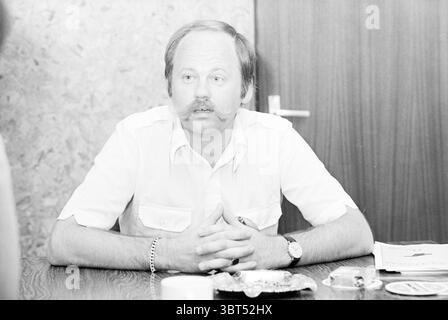Mr. Broek chairman Stormvogels Football people and trainers, Whizgle News, Dutch Desk, The Netherlands, 1950 - 2000 on 25-08-1981. These topics are shown in the image. In the scene, a middle-aged man sits at a wooden table, his hands clasped in front of him, conveying a sense of attentiveness. He has a well-groomed mustache and wears a light-colored, short-sleeved shirt that gives a casual yet professional appearance. His expression is serious and focused, suggesting an important conversation is taking place. Behind him, the walls are a textured tan, adding warmth to the environment, while a d Stock Photo