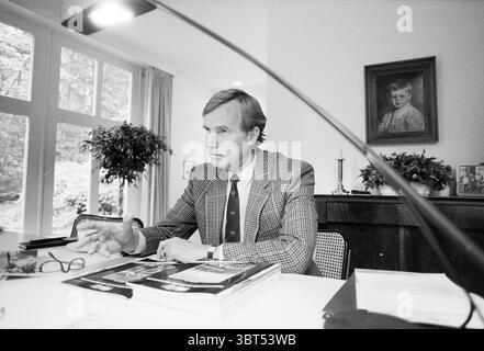 Heer Spoor Advertising agency, Whizgle News, Dutch Desk, The Netherlands, 1950 - 2000 on 24-09-1990. These are the elements in the image. The scene features a man seated at a sleek, modern desk, engaged in conversation. He has a thoughtful expression, his hand gesturing as he speaks. His attire is formal yet stylish, consisting of a patterned blazer that exudes sophistication. The workspace is thoughtfully arranged, with various magazines and documents piled neatly on the desk, suggesting an active engagement with work or ideas. A pair of glasses rests nearby, hinting at a moment of contemplat Stock Photo