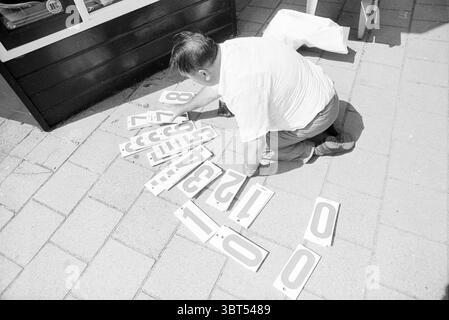 Select sticker letters and numbers, Whizgle News, Dutch Desk, The Netherlands, 1950 - 2000 in 1993. The image includes these topics. In a lively outdoor setting, a man is positioned on the ground, kneeling on a paved surface crafted from light-colored stones. He is focused intently on a collection of plastic numerals scattered around him. The numbers, painted in bold black on a white background, contrast sharply with the earthy tones of the ground. The man is wearing a short-sleeve, light-colored shirt that reflects a bright sunlight, suggesting a warm day. His hair is dark and slicked back, g Stock Photo
