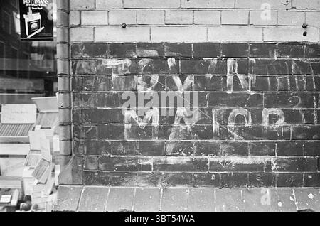 English text on wall Nassaulaan Muren Haarlem Nassaulaan The Netherlands, Whizgle News, Dutch Desk, The Netherlands, 1950 - 2000 on 31-01-1975. These topics are shown in the image. The scene depicts a textured brick wall, prominently featuring a faded, hand-painted inscription. The letters appear slightly irregular, suggesting they were applied with a rough brush or similar tool, and have softened over time, blending subtly into the wall's weathered surface. The bricks are mostly a dark gray, but the areas surrounding the inscription show signs of erosion, presenting various tones from deep ch Stock Photo